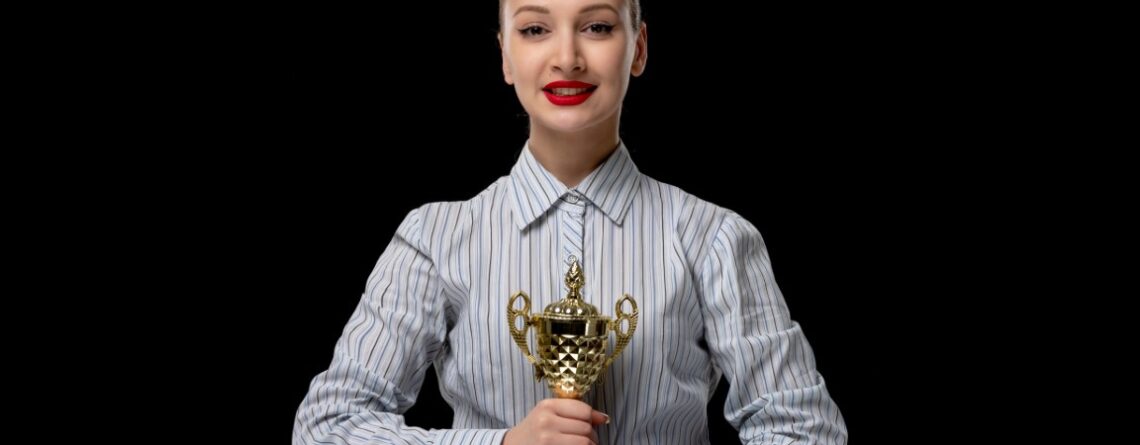 Image of a blonde businesswoman holding a golden trophy, dressed in an office costume and wearing red lipstick.